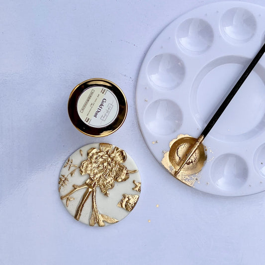 A small container of gold-colored edible baking decoration dust, alongside a cake with the dust applied to its surface, and a cake decorating brush with gold paint on it.