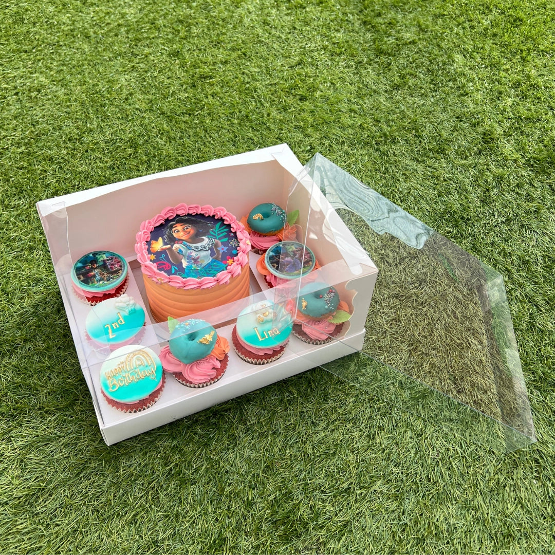 A clear cake box with a bento cake and eight cupcakes displayed in it, placed on a grassy surface.